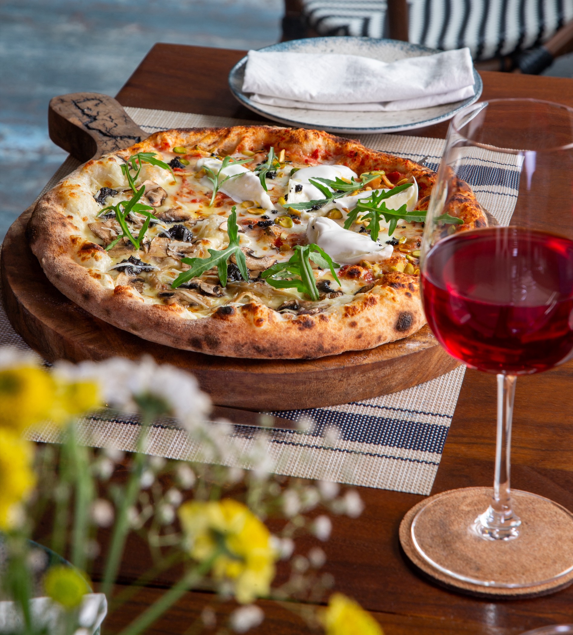 A wood-fired pizza with mushrooms, burrata and rocket beside a glass of red wine on a sunlit table at Rosmarino, Indiranagar.