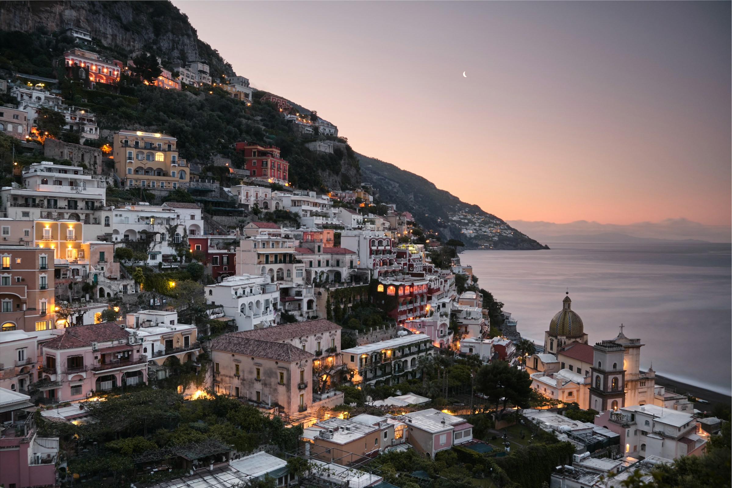 Positano on the Amalfi Coast at dusk — pastel cliffside houses glowing with warm light above the Mediterranean